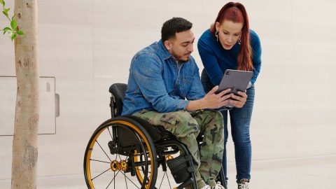 Man in a wheelchair holding tablet to share with a woman standing nearby