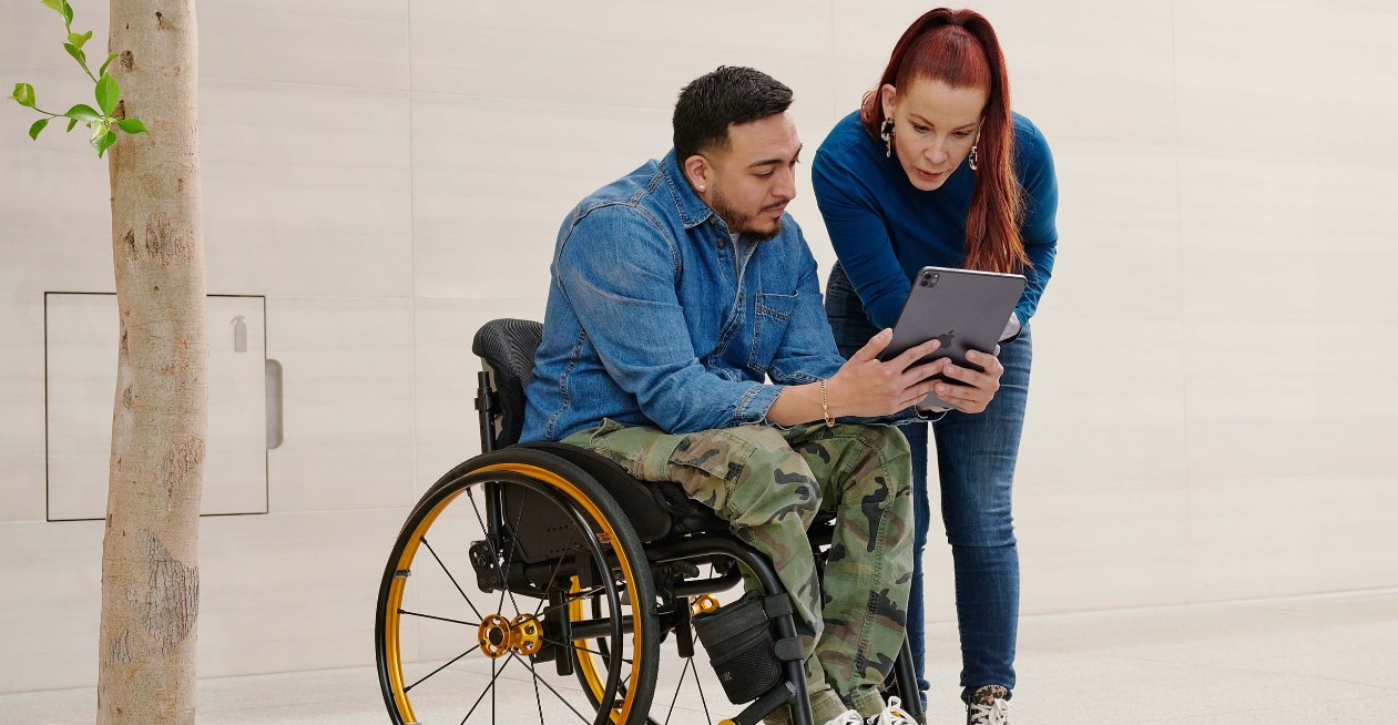 Man in a wheelchair holding tablet to share with a woman standing nearby