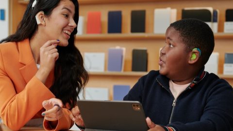 A woman wearing a hearing aid showing a boy wearing a hearing aid how to use an iPad.